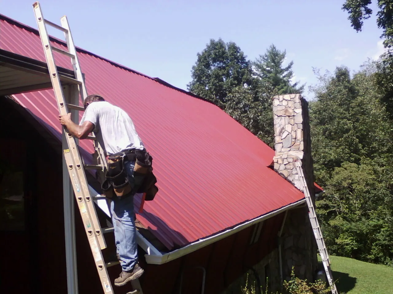 Roofer on a ladder beside a red metal roof and stone chimney — Soffit & Fascia Repair by Roofing Brothers in Arriba